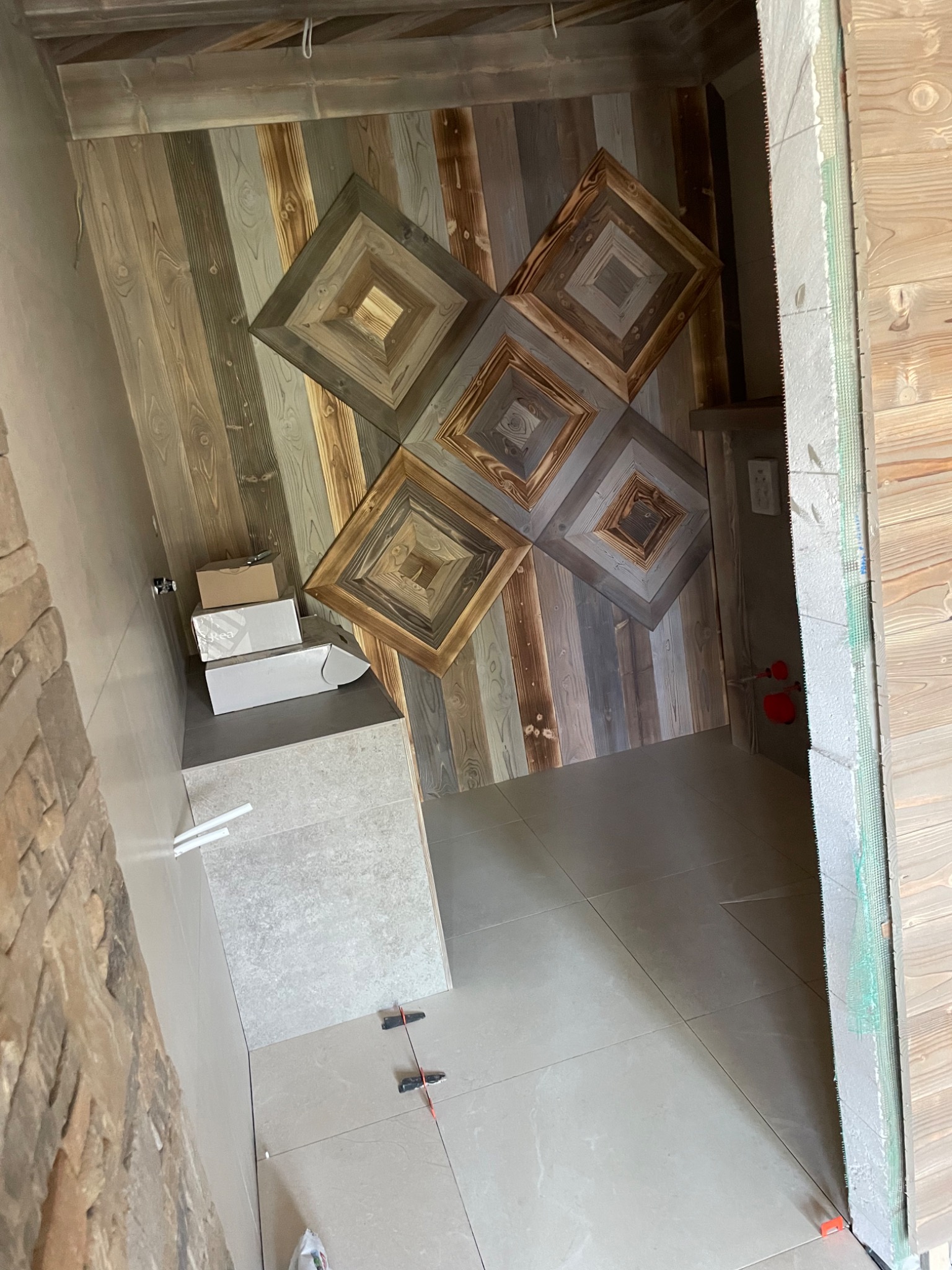 Wnętrze łazienki w trakcie prac wykończeniowych; widoczne drewniane panele ścienne z geometrycznym wzorem, jasne płytki podłogowe z krzyżykami dystansowymi, kartony na blacie i fragment ściany...