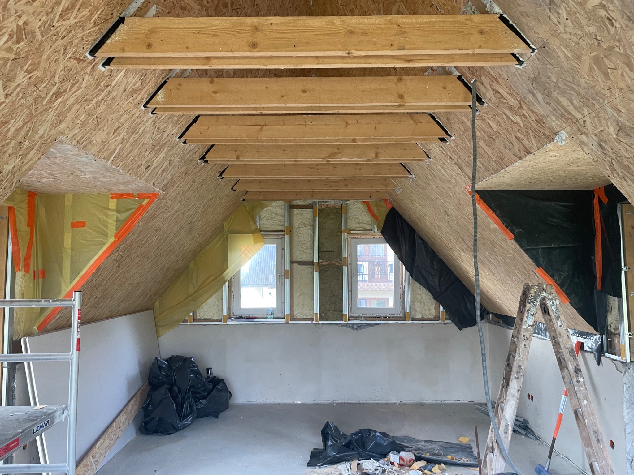 Poddasze w trakcie adaptacji: widoczne drewniane belki stropowe z czarnymi elementami mocującymi, ściany obłożone płytami OSB, okna z izolacją, częściowo otynkowana ściana, worki z odpadami...