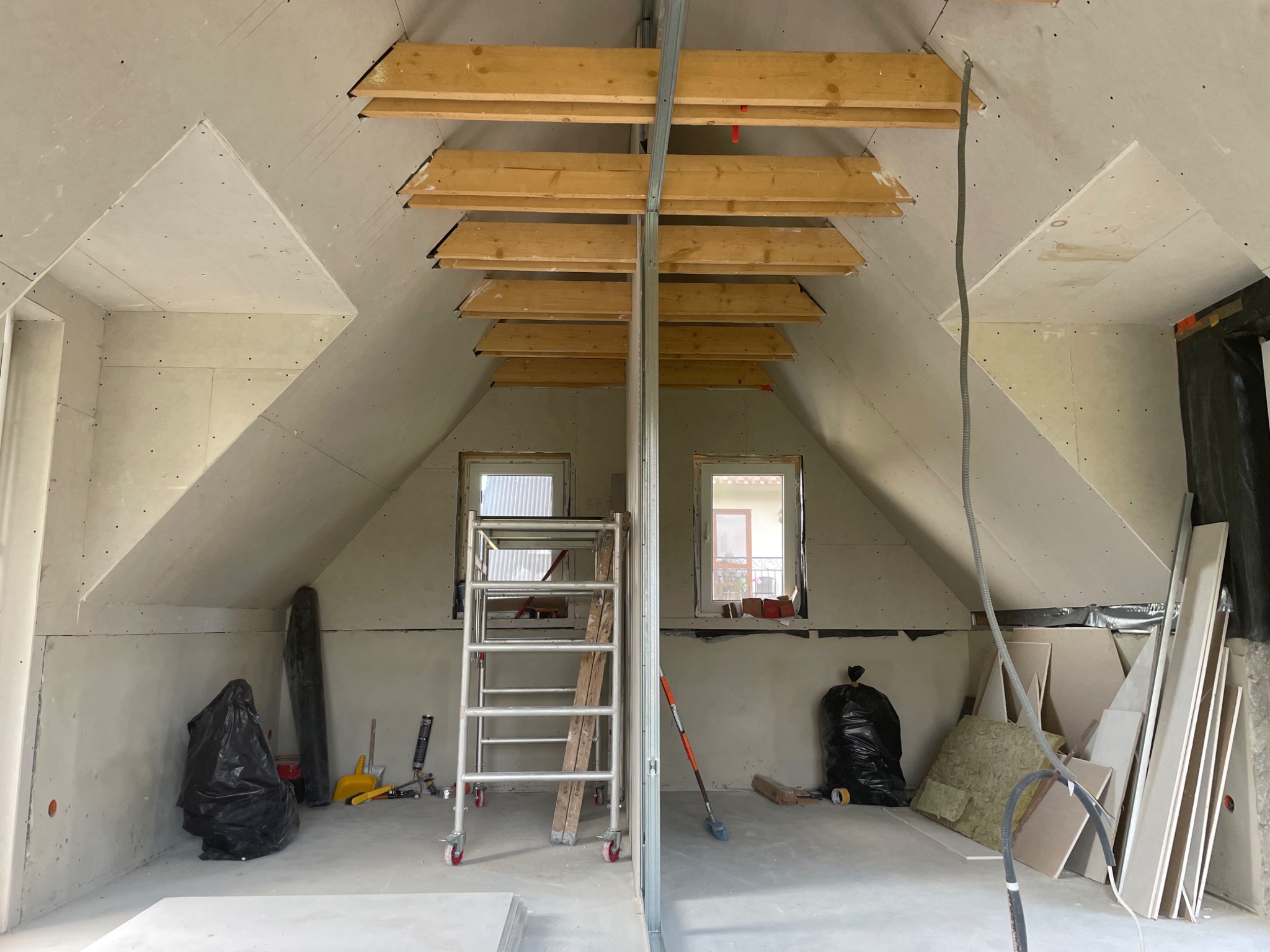 Poddasze w trakcie adaptacji w Bukowinie Tatrzańskiej: widoczne drewniane elementy konstrukcyjne pod sufitem, ściany obłożone płytami gipsowo-kartonowymi, drabina aluminiowa i materiały budowlane...