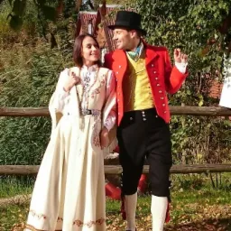 Para w tradycyjnych strojach ludowych: kobieta w kremowej sukni z haftowanymi detalami i srebrnym pasem, mężczyzna w czerwonej marynarce, żółtej kamizelce, czarnych spodniach i cylindrze, stojąca...