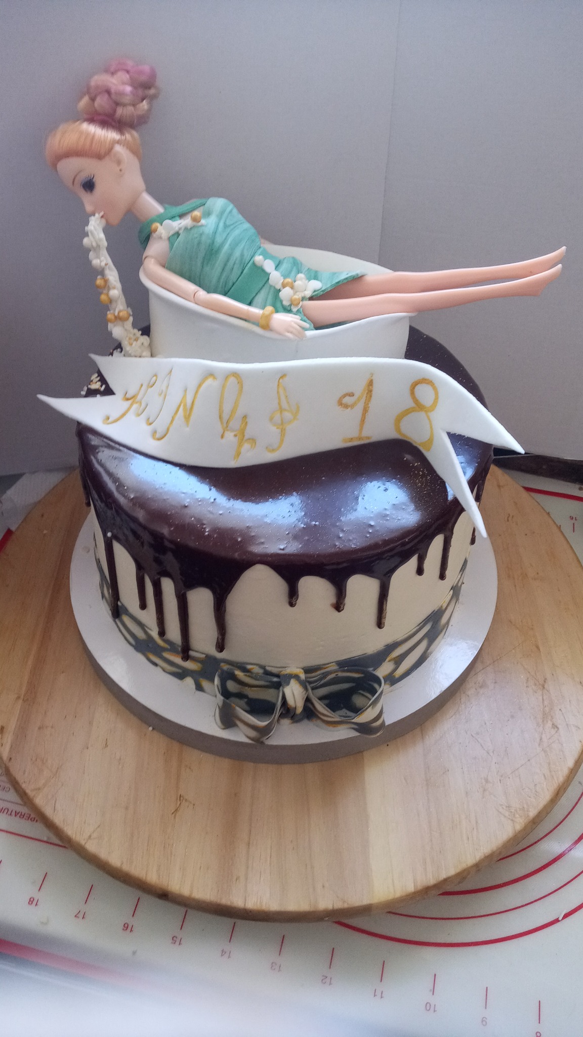 Dwupoziomowy tort urodzinowy z polewą czekoladową i dekoracją w postaci lalki w kąpieli, z napisem 'Kinga 18' na białej wstążce.