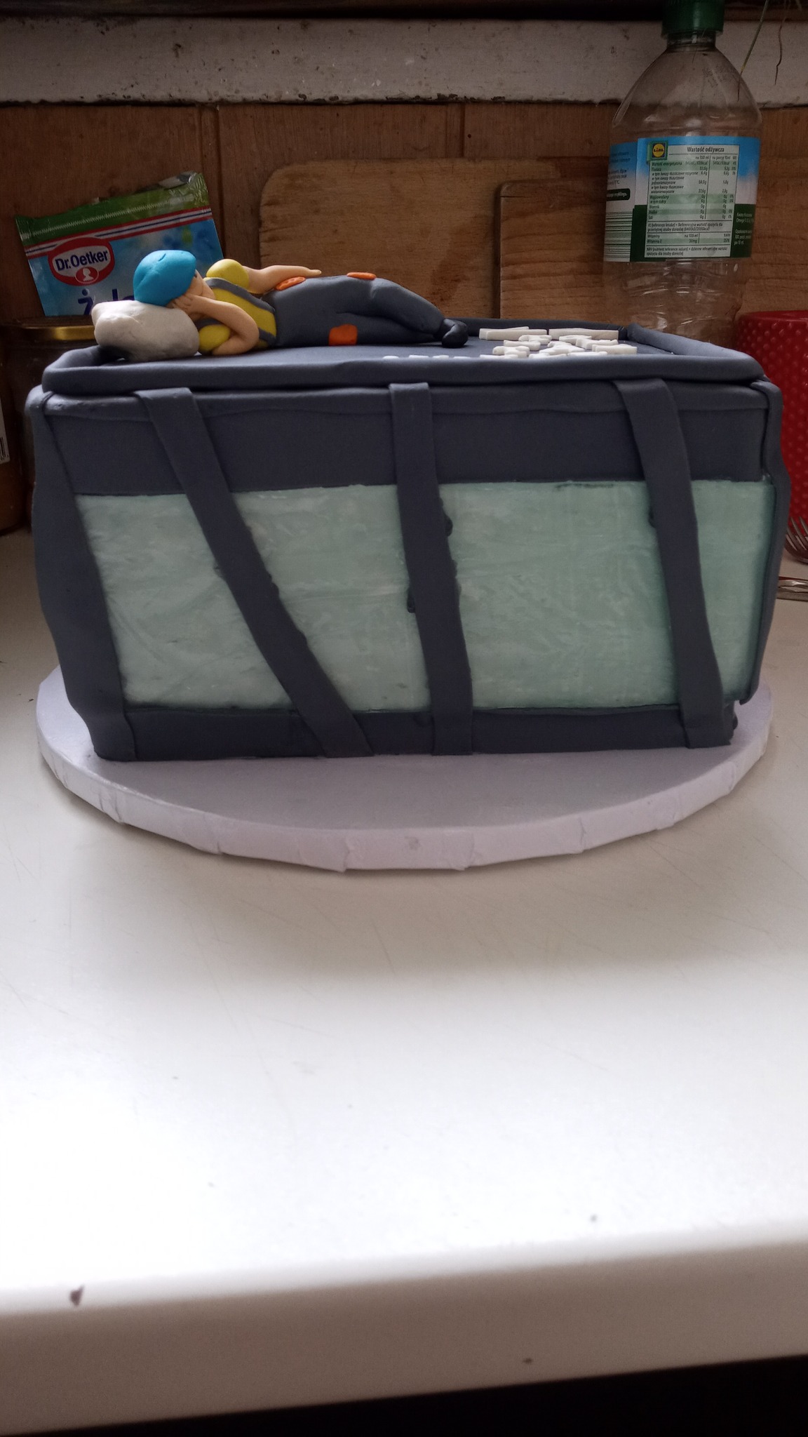 Tort w kształcie skrzyni z figurką śpiącego mężczyzny na górze, wykonany z masy cukrowej, umieszczony na białym podkładzie, z widocznymi produktami spożywczymi w tle.