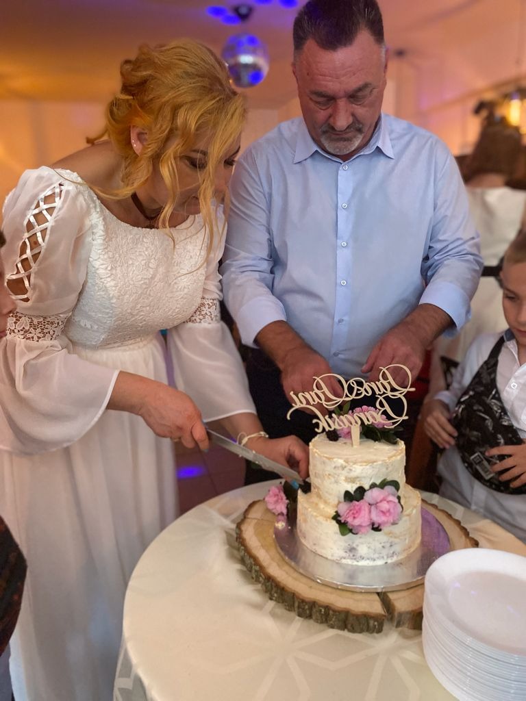 Para młoda kroi dwupiętrowy tort weselny z jasnym kremem i różowymi kwiatami, umieszczony na drewnianym plastrze, podczas uroczystości.