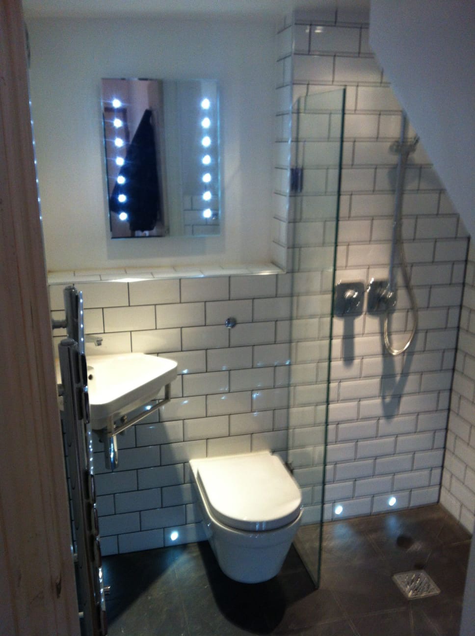 Nowoczesna łazienka z białymi, prostokątnymi płytkami, przeszklonym prysznicem, podświetlanym lustrem i punktowym oświetleniem podłogowym