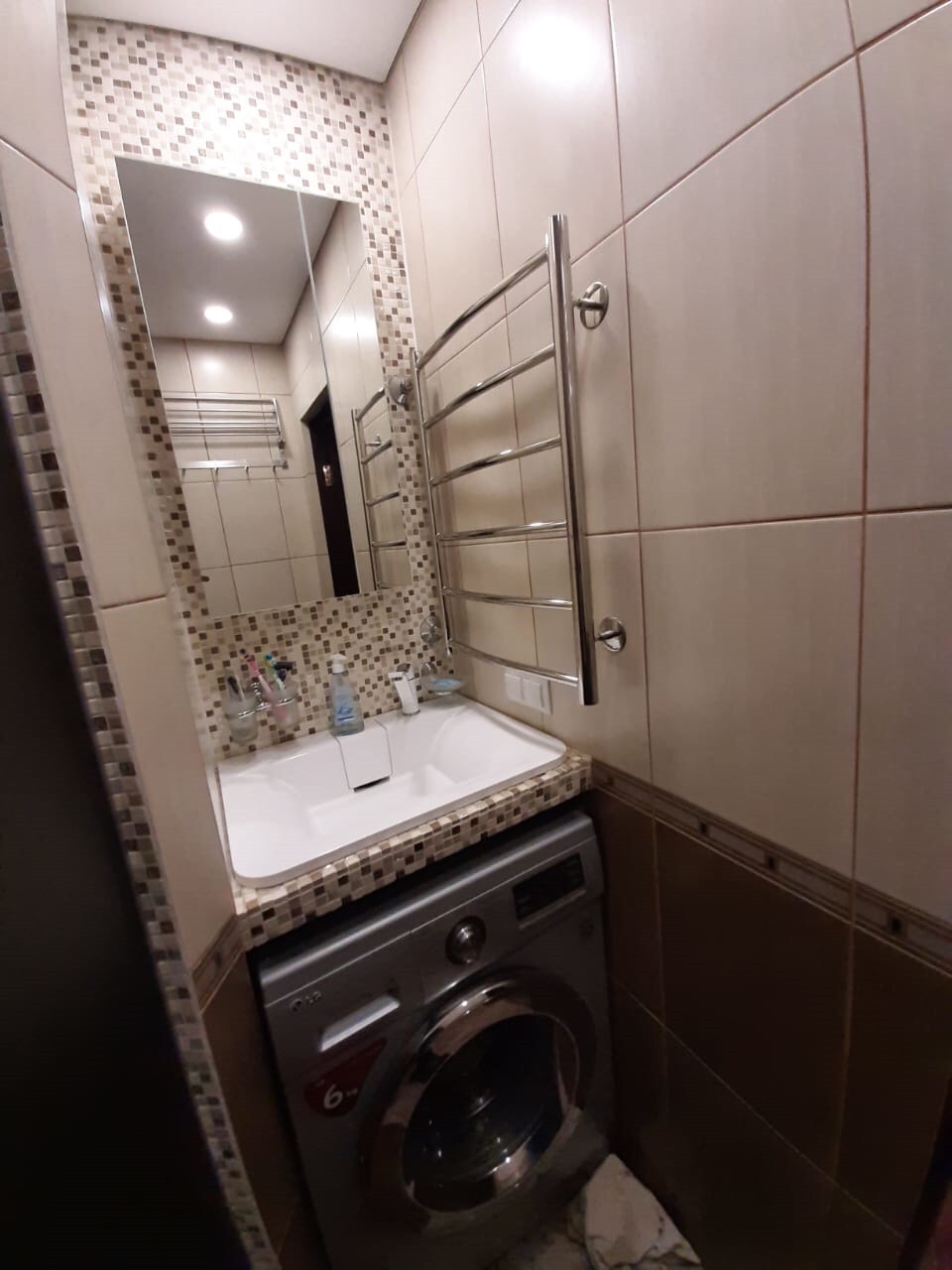 Kompaktowa łazienka z umywalką nad pralką, mozaikowa ściana i grzejnik drabinkowy, widoczne kosmetyki i akcesoria.