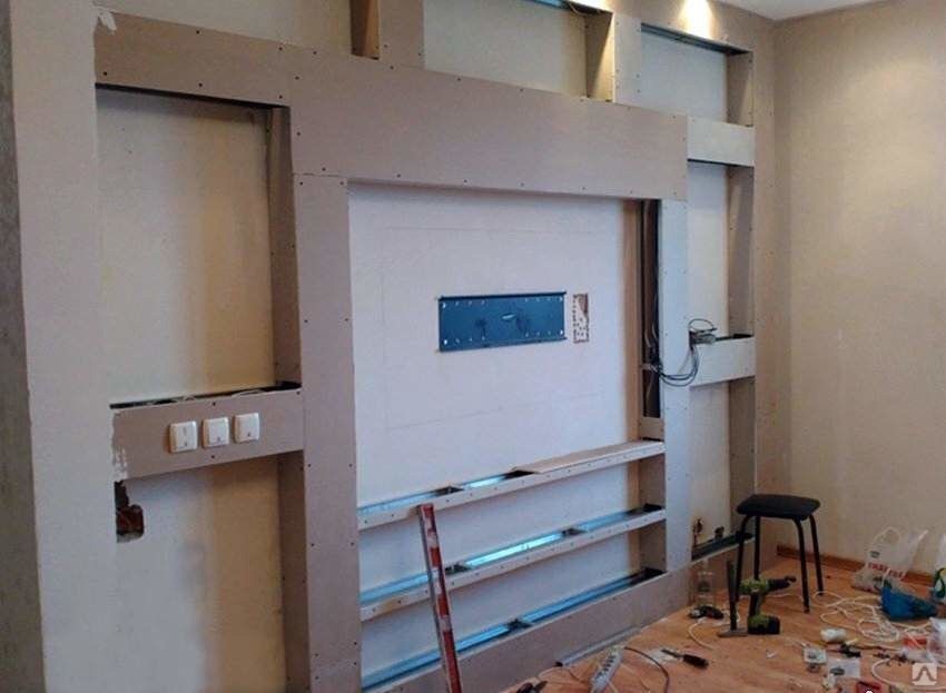 Ściana w trakcie zabudowy z płyt kartonowo-gipsowych, widoczne metalowe profile, puszka instalacyjna i fragment instalacji elektrycznej, na podłodze narzędzia i stołek.