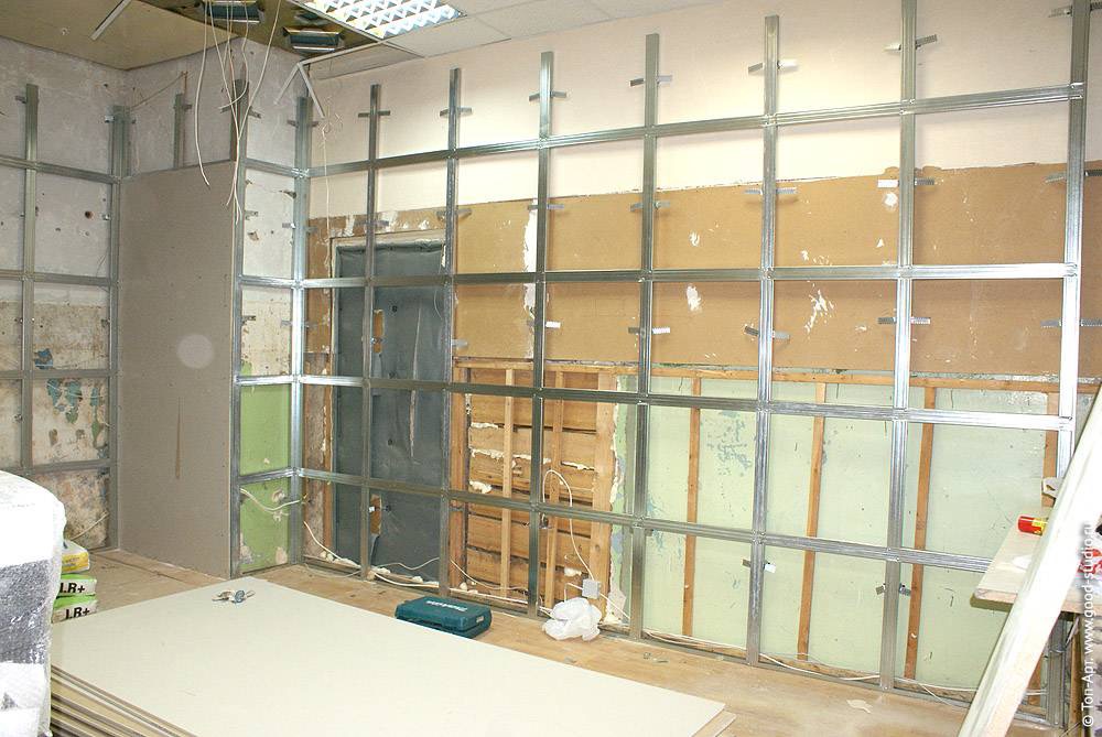 Montaż metalowego stelażu pod okładzinę z płyt na ścianie w trakcie prac wykończeniowych, widoczne kable i narzędzia, częściowo zamontowane płyty.