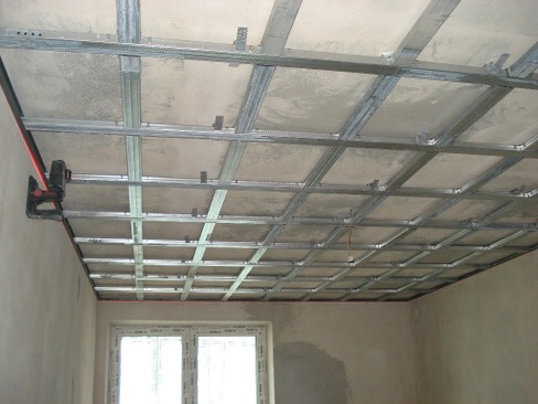 Montaż metalowej konstrukcji sufitu podwieszanego z płyt kartonowo-gipsowych, widoczny laserowy niwelator na ścianie, surowy stan wykończenia wnętrza.