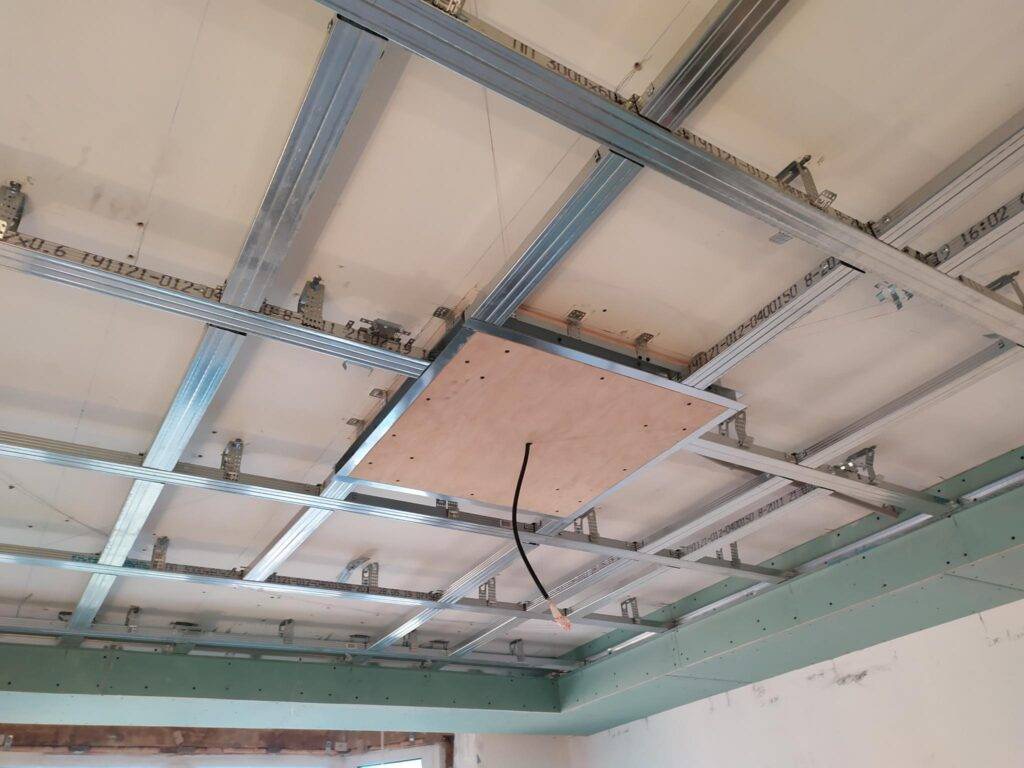 Stelaż sufitu podwieszanego z płytami kartonowo-gipsowymi w trakcie montażu, widoczne profile metalowe, płyty oraz otwór rewizyjny z wystającym przewodem elektrycznym.