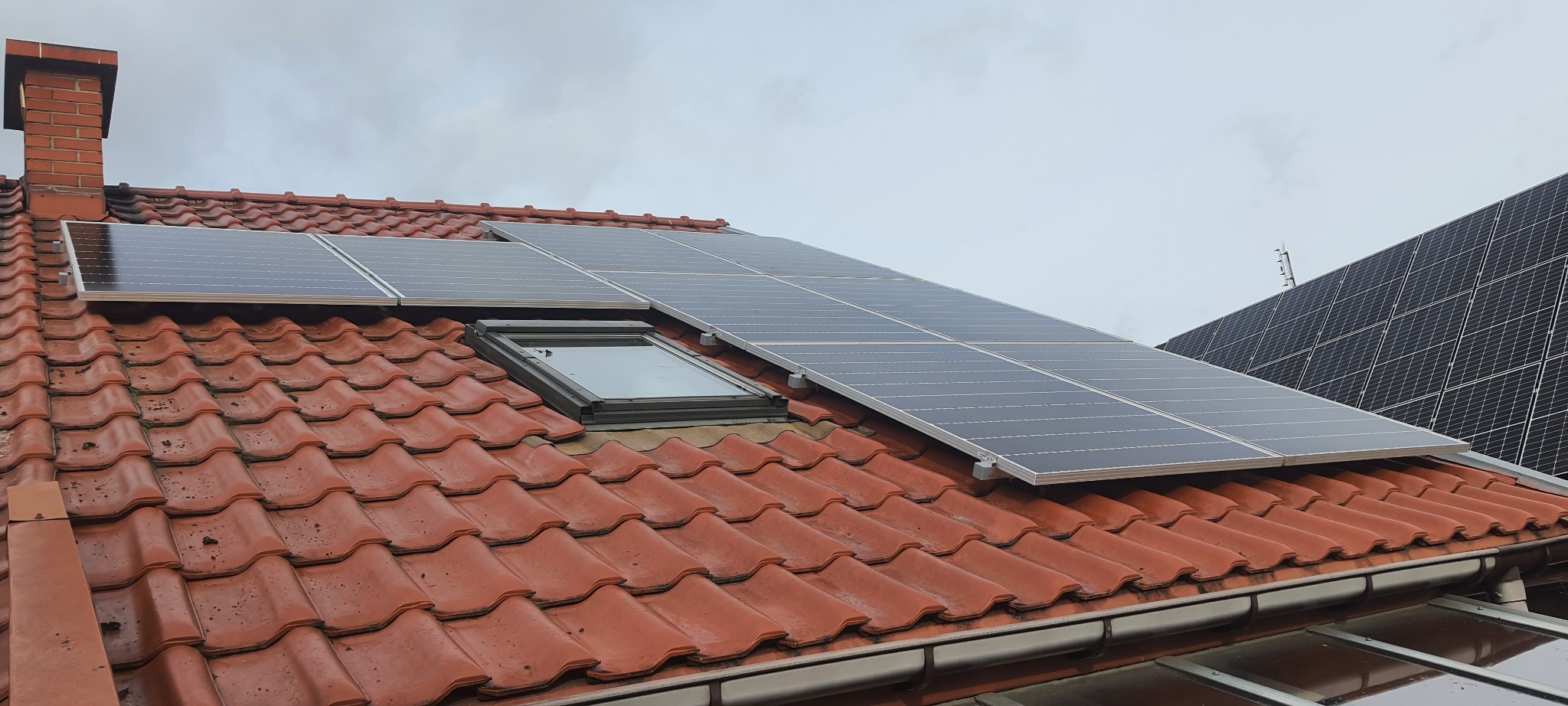 Czerwona dachówka z zamontowanymi panelami słonecznymi obok okna dachowego i ceglanego komina, dodatkowe panele widoczne w tle.