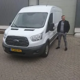 Biały samochód dostawczy Ford Transit z kierowcą stojącym obok na tle szarego magazynu z bramą rolowaną