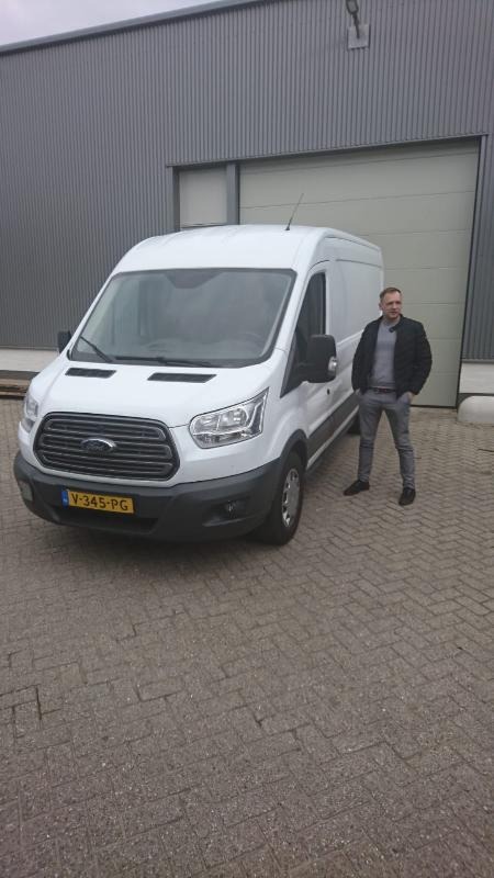 Biały samochód dostawczy Ford Transit z kierowcą stojącym obok na tle szarego magazynu z bramą rolowaną