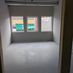 Firma ogólnobudowlana SZWARCBUD - Surowe wnętrze w trakcie wykańczania: szare ściany z płyt gipsowych, betonowa podłoga, trzy okna z widokiem na kontener i drzewa, widoczne rury wentylacyjne pod sufitem.