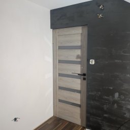 Firma ogólnobudowlana SZWARCBUD - Wnętrze w trakcie prac wykończeniowych: zamontowane drzwi wewnętrzne z przeszkleniami, ściana pomalowana na ciemny szary kolor z efektem dekoracyjnym, druga ściana biała, widoczne gniazdka...