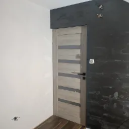 Wnętrze w trakcie prac wykończeniowych: zamontowane drzwi wewnętrzne z przeszkleniami, ściana pomalowana na ciemny szary kolor z efektem dekoracyjnym, druga ściana biała, widoczne gniazdka...