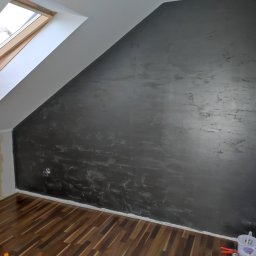 Firma ogólnobudowlana SZWARCBUD - Ściana w pokoju na poddaszu, pomalowana na ciemny kolor z efektem dekoracyjnym, widoczne narzędzia malarskie i drewniana podłoga.