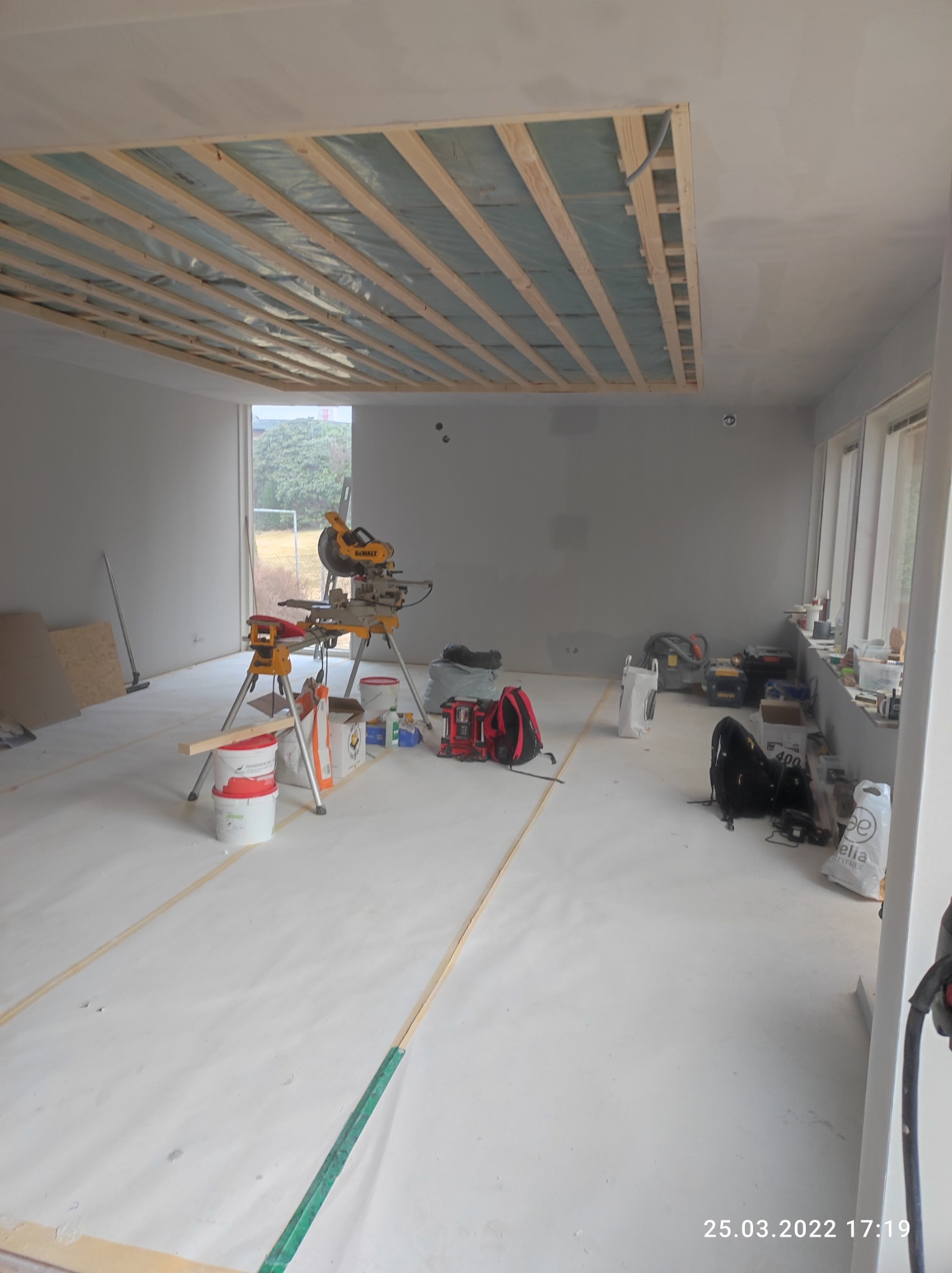 Wnętrze w trakcie prac remontowych: widoczna konstrukcja drewniana podwieszanego sufitu, zabezpieczona folią, narzędzia budowlane, biała folia ochronna na podłodze, ściany przygotowane do malowania.