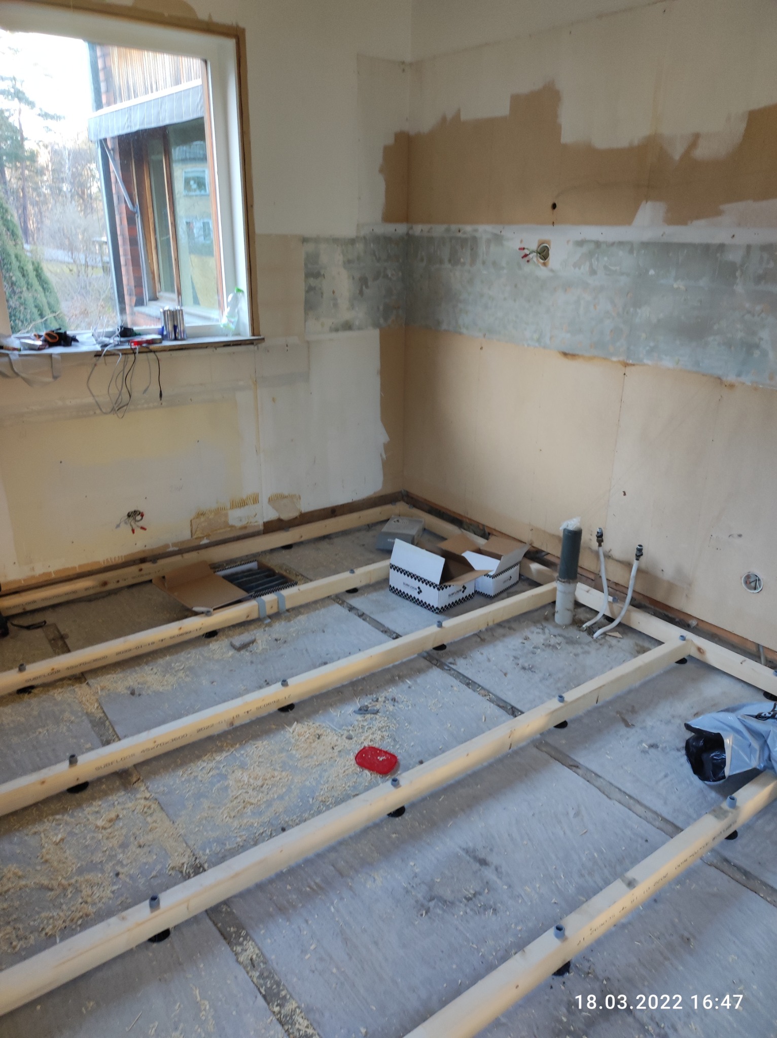Wnętrze w trakcie remontu: widoczne drewniane legary pod podłogę, odkryte fragmenty ścian, instalacje hydrauliczne i elektryczne bez wykończenia oraz okno z widokiem na zieleń.