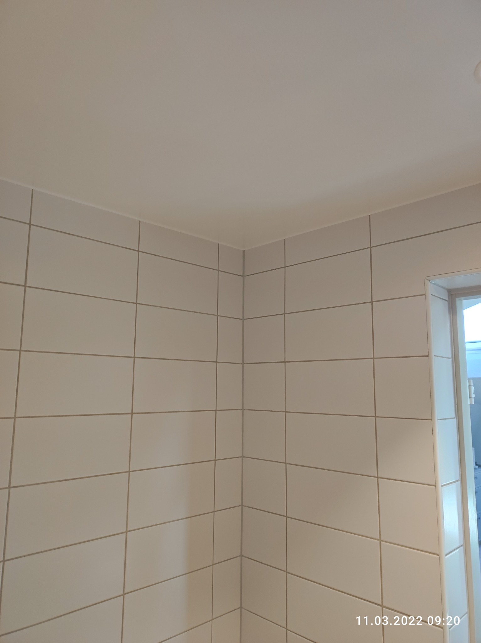 Świeżo wykafelkowana ściana w łazience, z widocznym narożnikiem i fragmentem ościeżnicy drzwiowej, wykonana z białych, prostokątnych płytek z szarą fugą.