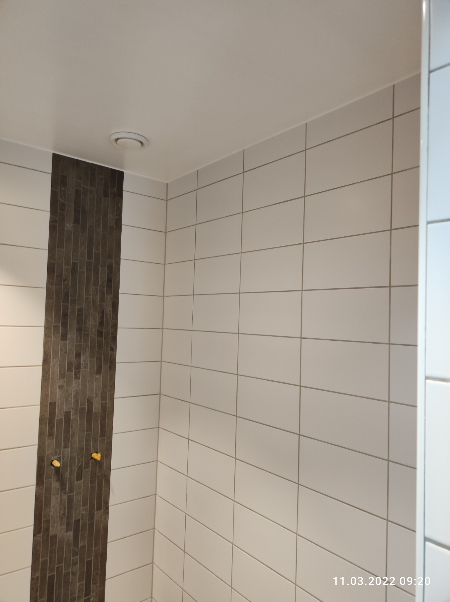 Wykończona łazienka z białymi płytkami i pionowym pasem ciemnych, cegiełkowych płytek, widoczne dwa żółte zaślepki na rury, sufit z okrągłym nawiewnikiem.