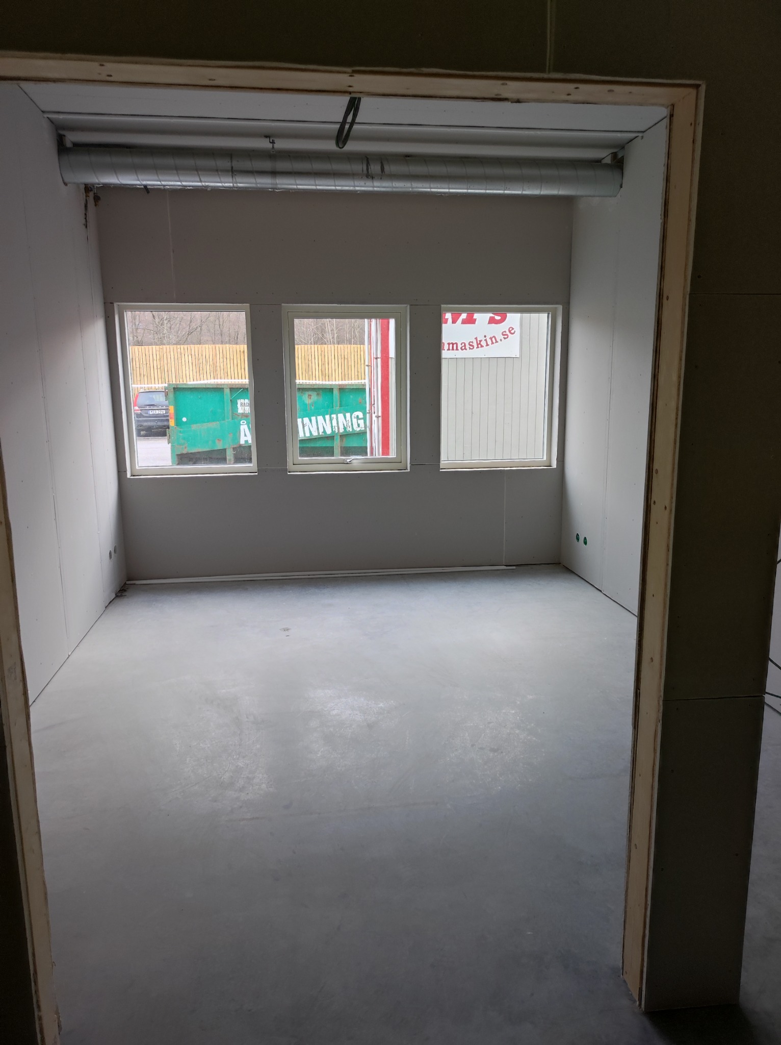 Surowe wnętrze w trakcie wykańczania: szare ściany z płyt gipsowych, betonowa podłoga, trzy okna z widokiem na kontener i drzewa, widoczne rury wentylacyjne pod sufitem.