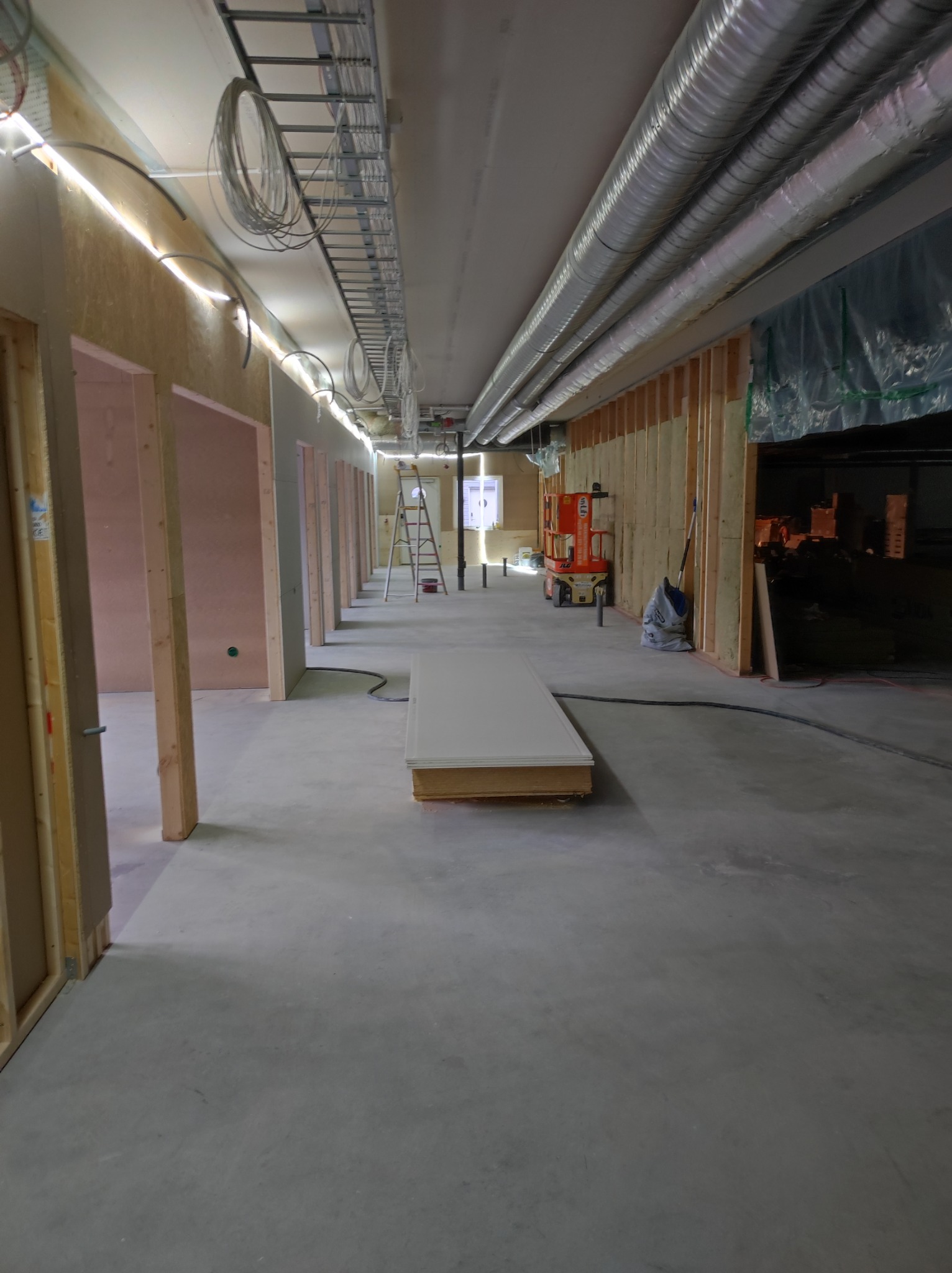 Wnętrze w trakcie prac wykończeniowych, widoczne ramy ścianek działowych, instalacje pod sufitem, podnośnik nożycowy i materiały budowlane w tle.