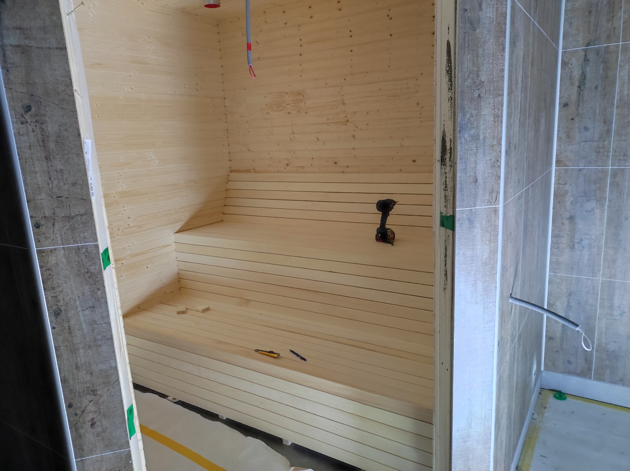 Wnętrze sauny w trakcie budowy: drewniane deski, ławy, wkrętarka i nożyk na ławie, widoczne kable elektryczne pod sufitem, płytki na ścianie obok.