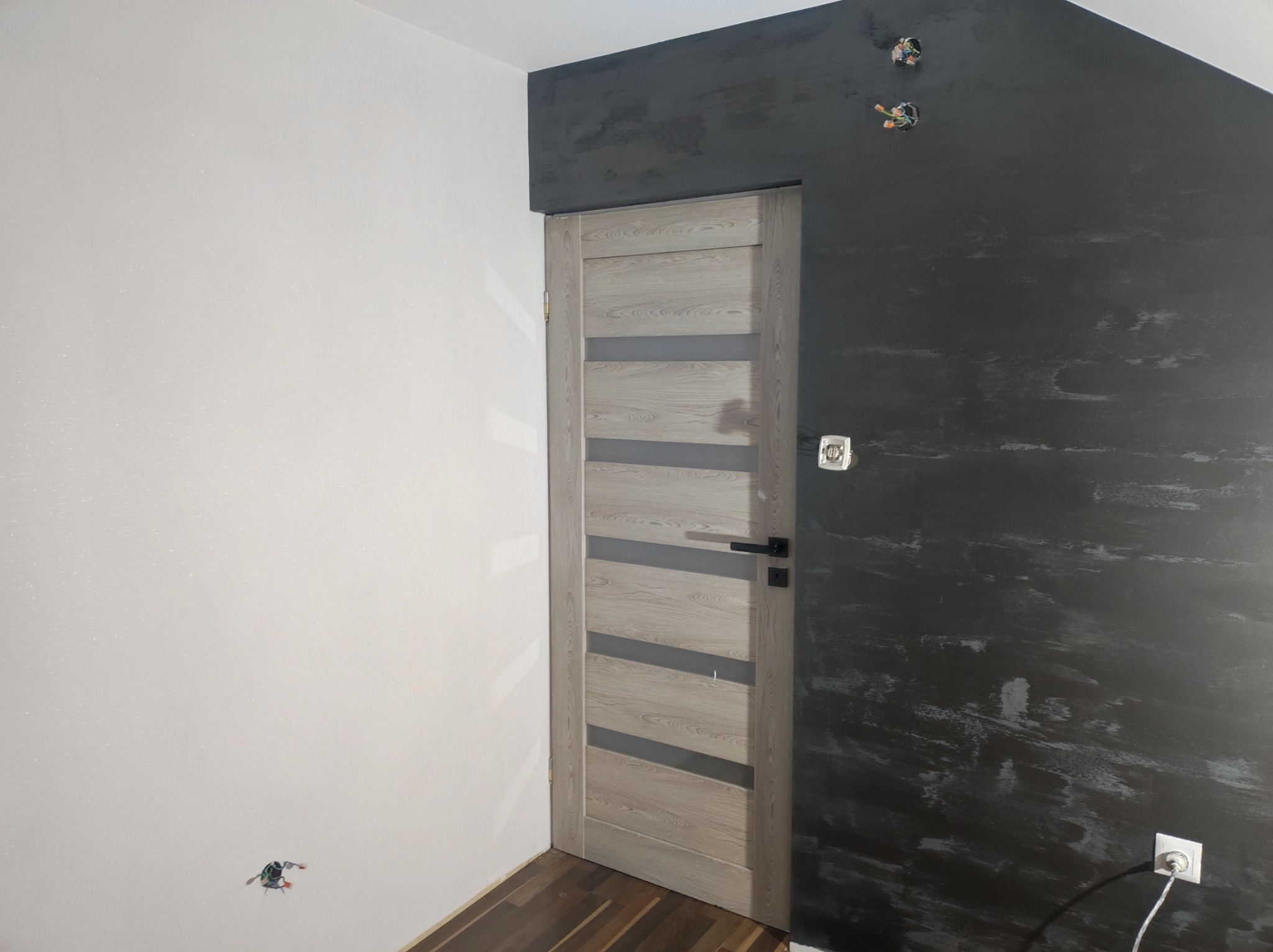 Wnętrze w trakcie prac wykończeniowych: zamontowane drzwi wewnętrzne z przeszkleniami, ściana pomalowana na ciemny szary kolor z efektem dekoracyjnym, druga ściana biała, widoczne gniazdka...