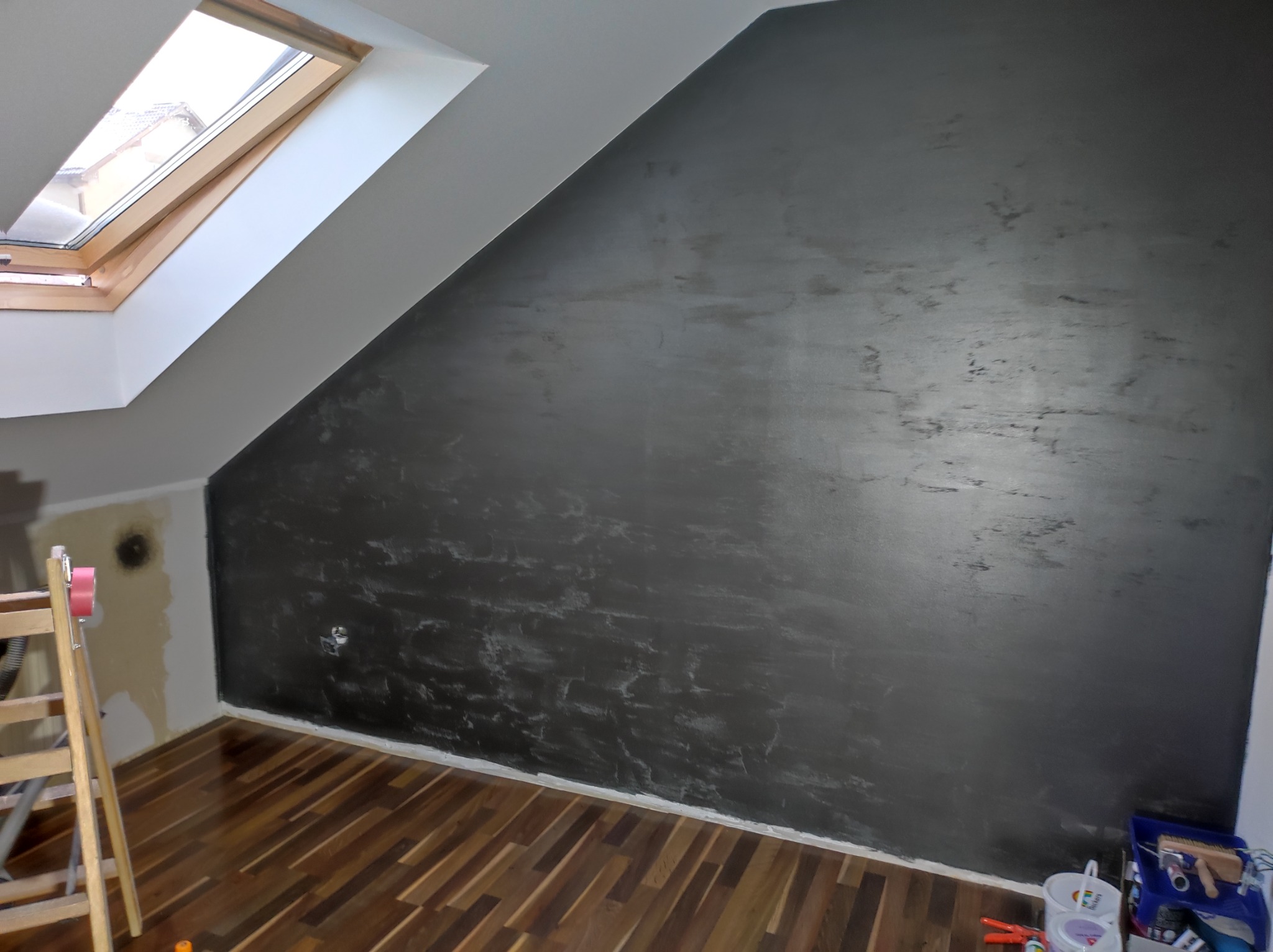 Ściana w pokoju na poddaszu, pomalowana na ciemny kolor z efektem dekoracyjnym, widoczne narzędzia malarskie i drewniana podłoga.