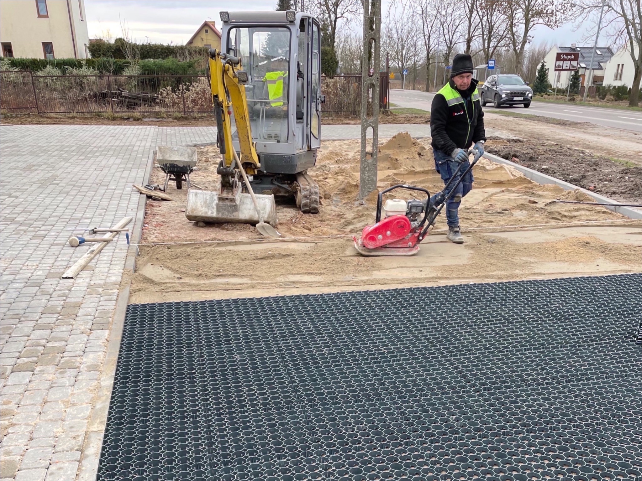 Pracownik zagęszcza piasek pod podbudowę z kratki trawnikowej, obok stoi mini-koparka i ułożona kostka brukowa, widoczna droga i zabudowania w tle.