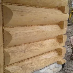 Fragment ściany zewnętrznej budynku z bali drewnianych, z dekoracyjnymi elementami wykończeniowymi, widoczne kamienne fundamenty i schody betonowe.