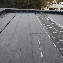 Widok z góry na płaski dach pokryty papą termozgrzewalną z widocznymi pasami z logotypem IZOBUD, tło stanowią korony drzew i fragment budynku w stanie surowym.