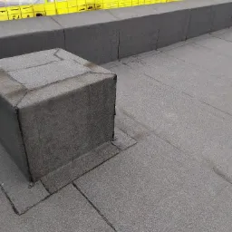 Szczegółowe ujęcie płaskiego dachu pokrytego papą termozgrzewalną, z widocznym kominkiem wentylacyjnym i fragmentem zabezpieczenia krawędzi dachu w tle.