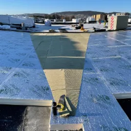 Montaż izolacji termicznej Kingspan Therma na dachu płaskim, widoczne narzędzia pomiarowe i częściowo ułożona warstwa wełny mineralnej, w tle zabudowa miejska pod błękitnym niebem.