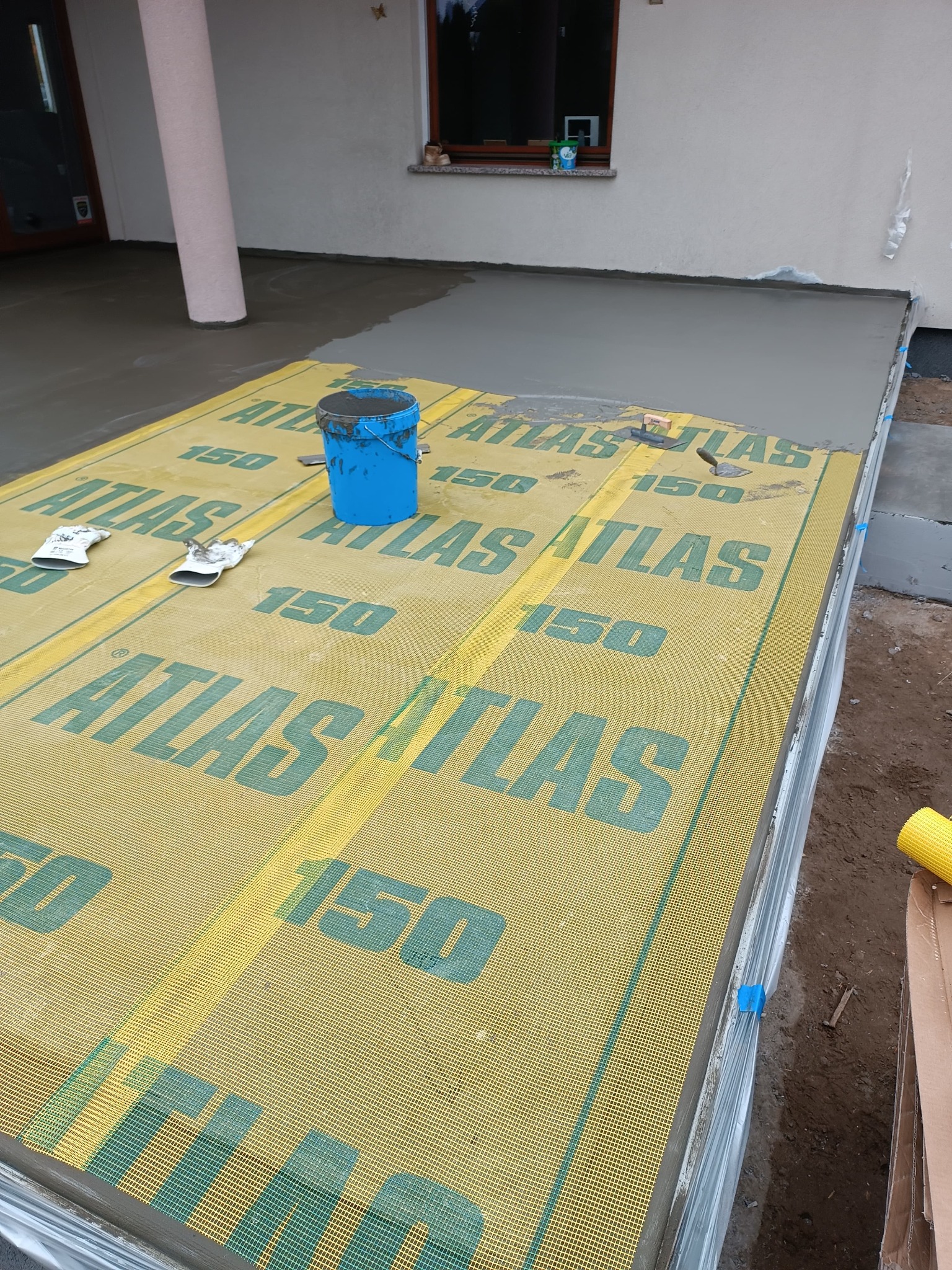 Świeżo wylana, szara wylewka na zewnątrz budynku, zabezpieczona żółtą folią budowlaną z zielonym nadrukiem 'ATLAS 150'. Widoczne narzędzia, wiadro i rękawice robocze.