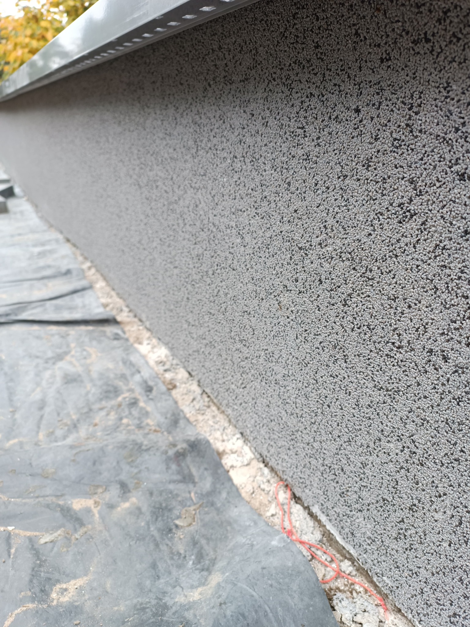 Szara ściana z fakturą baranka, zabezpieczona czarną folią i listwą okapową, widoczny fragment czerwonego sznurka.