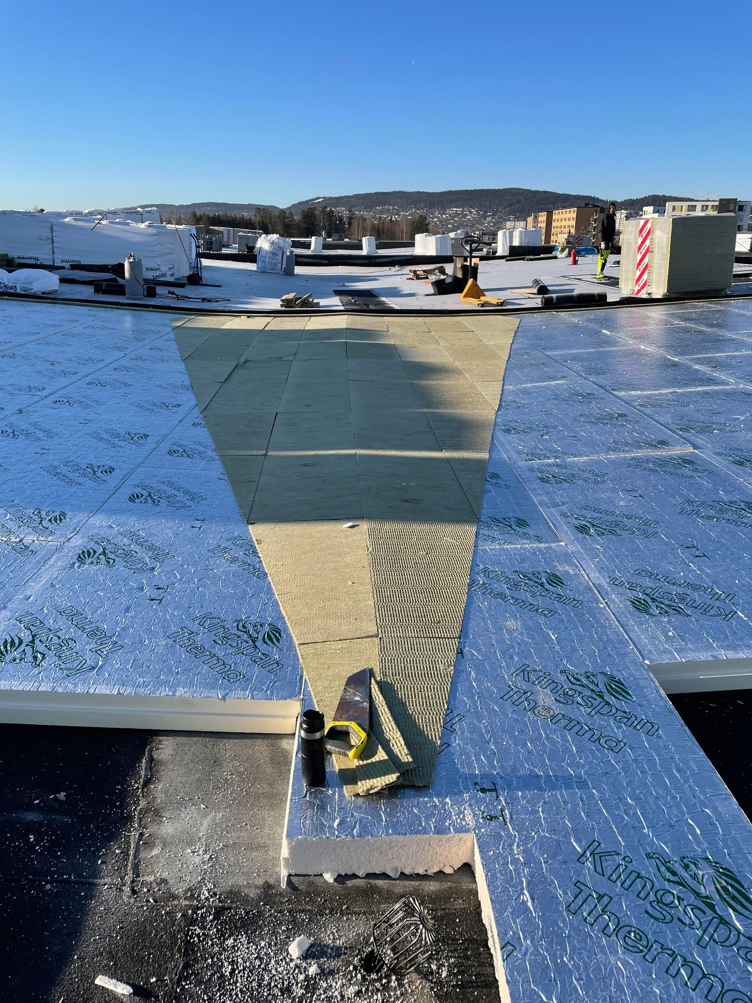 Montaż izolacji termicznej Kingspan Therma na dachu płaskim, widoczne narzędzia pomiarowe i częściowo ułożona warstwa wełny mineralnej, w tle zabudowa miejska pod błękitnym niebem.