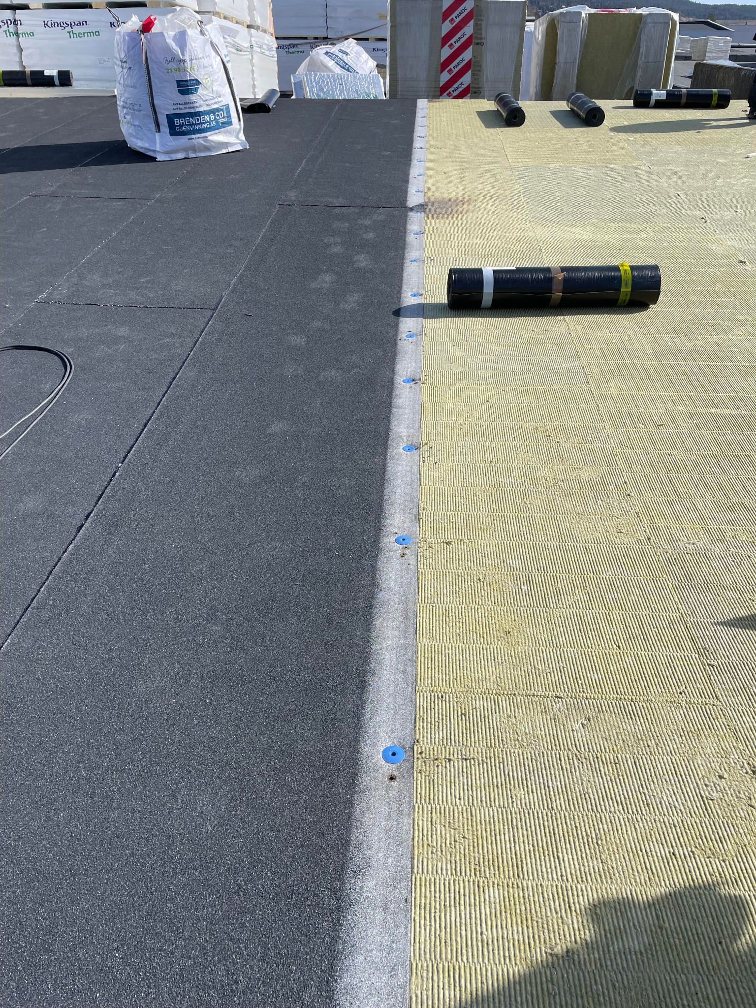 Fragment dachu w trakcie prac: z jednej strony ułożona czarna papa, z drugiej żółta wełna mineralna, widoczne rolki papy i worki z materiałami izolacyjnymi.