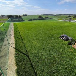 KADOKA Krzysztof Kamiński - Automatyczna kosiarka parkuje przy ceglanym murku na tle idealnie przystrzyżonego trawnika z widokiem na pola.