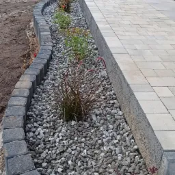 Krawężnik z kostki obramowujący rabatę z szarym grysem, ozdobne trawy i krzewy, przylegający do jasnej kostki brukowej tarasu.