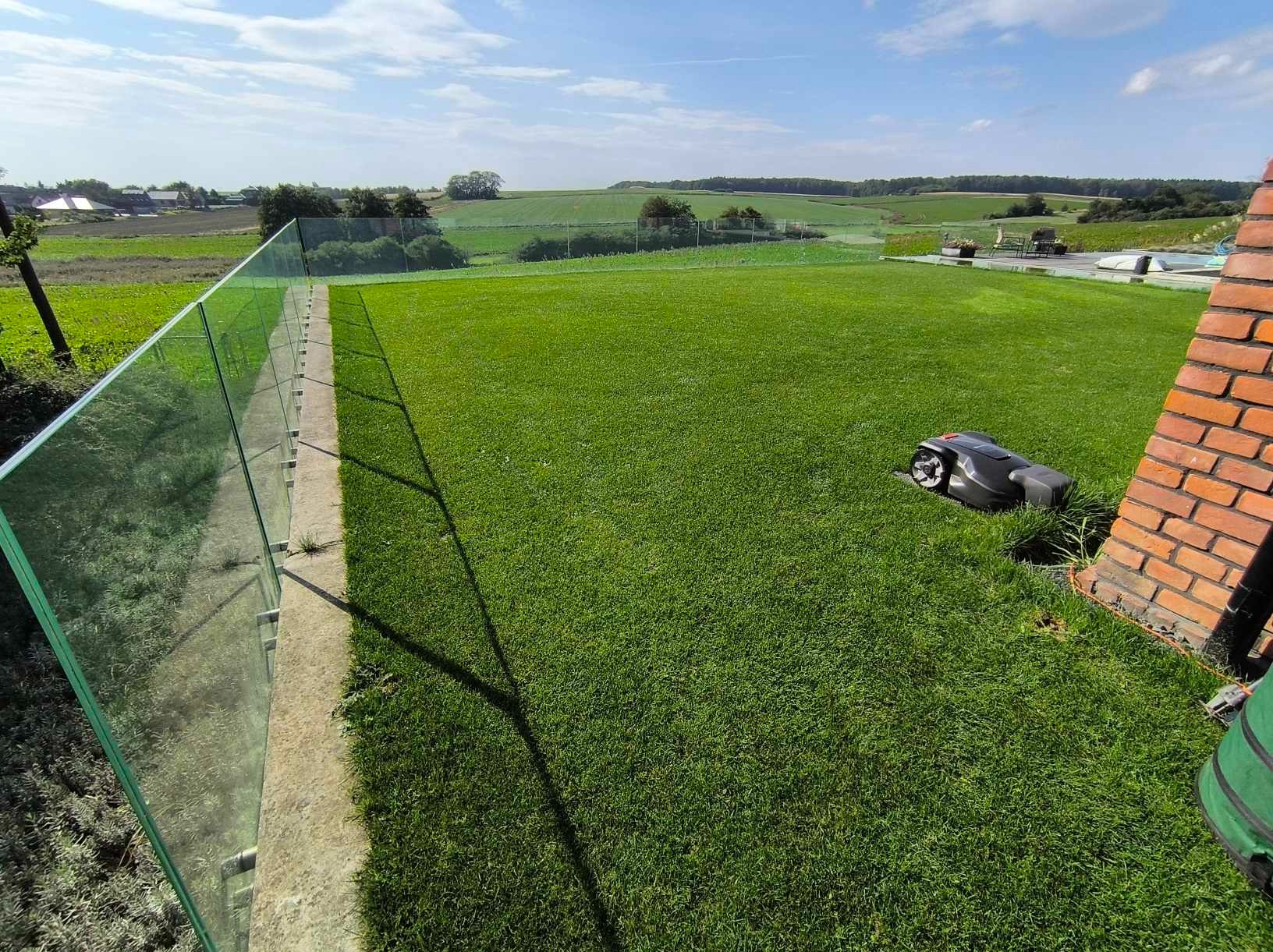 Automatyczna kosiarka parkuje przy ceglanym murku na tle idealnie przystrzyżonego trawnika z widokiem na pola.
