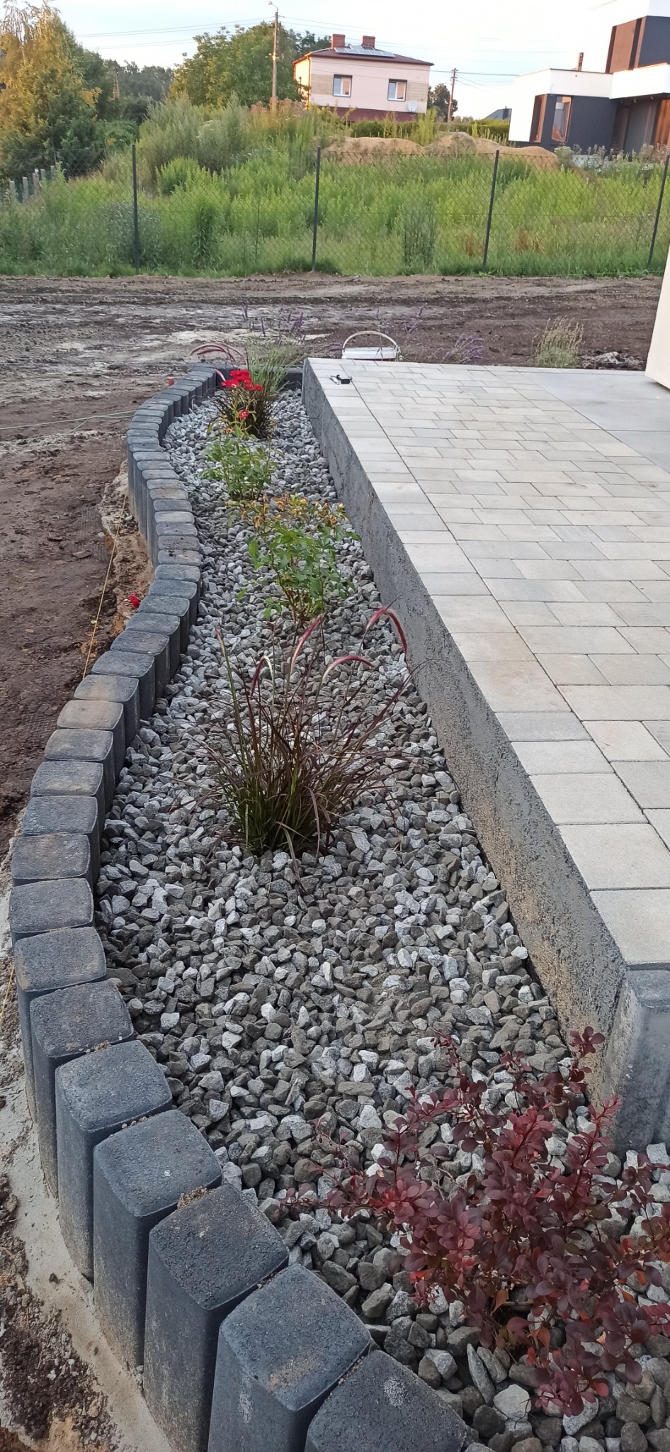 Krawężnik z kostki obramowujący rabatę z szarym grysem, ozdobne trawy i krzewy, przylegający do jasnej kostki brukowej tarasu.