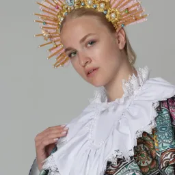 Portret młodej kobiety w stylizowanej sukni i bogato zdobionym, różowo-złotym nakryciu głowy z perłami i kameą, pozującej na jasnoszarym tle.