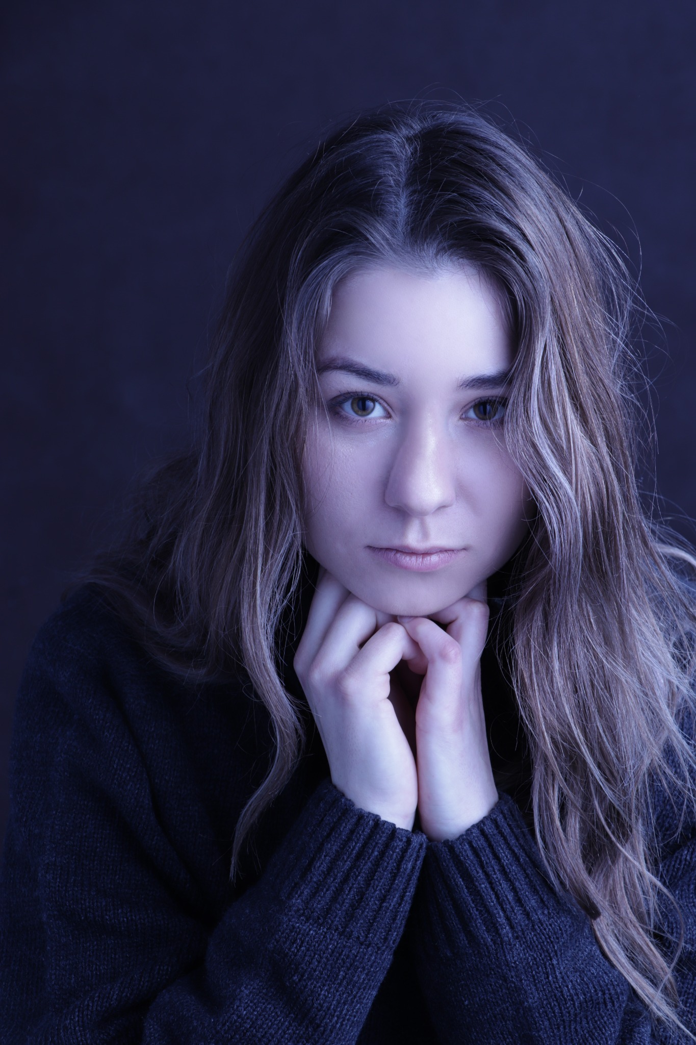 Portret młodej kobiety z długimi, blond włosami i zielonymi oczami, opierającej brodę na dłoniach, na ciemnym, rozmytym tle.