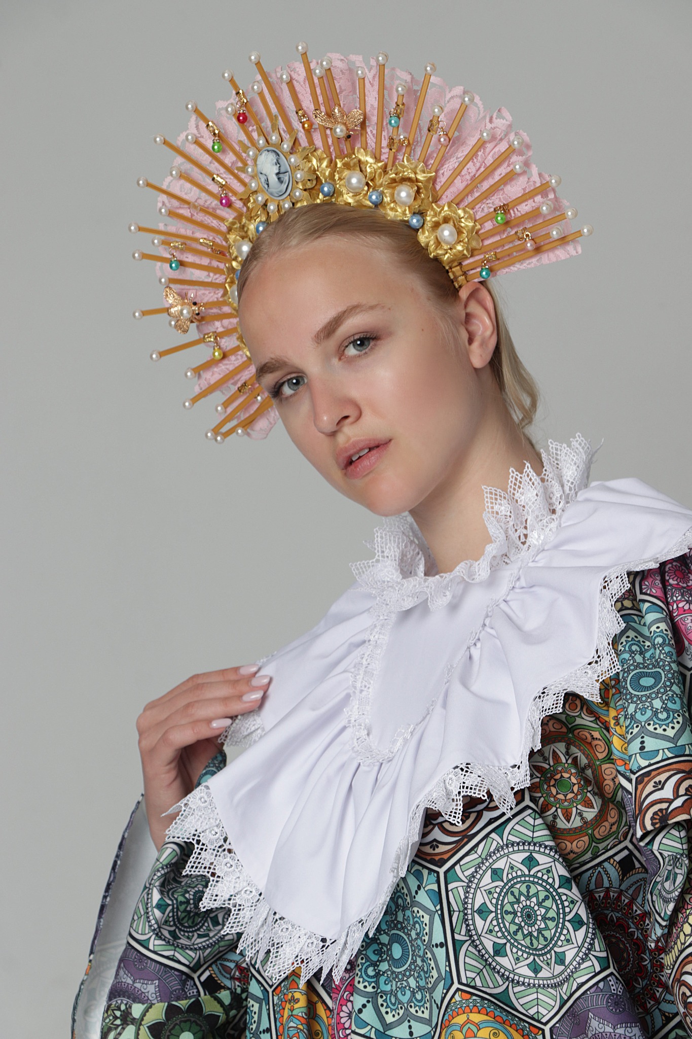 Portret młodej kobiety w stylizowanej sukni i bogato zdobionym, różowo-złotym nakryciu głowy z perłami i kameą, pozującej na jasnoszarym tle.