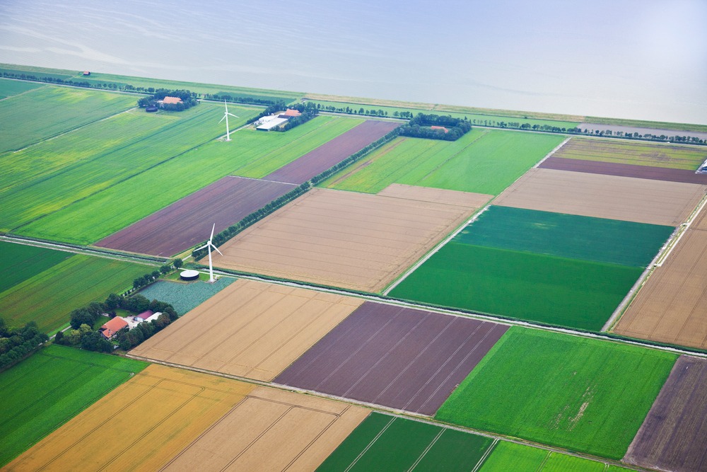 Widok z lotu ptaka na mozaikę pól uprawnych o różnych kolorach i teksturach, z farmami i turbinami wiatrowymi w tle, oddzielonych drogami i kanałami.