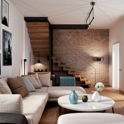 Magdalena Olejniczak Projektowanie Wnętrz - Jasny salon z beżową sofą, drewnianymi schodami z ceglaną ścianą w tle, okrągłym białym stolikiem kawowym z wazonami oraz galerią obrazów na ścianie.