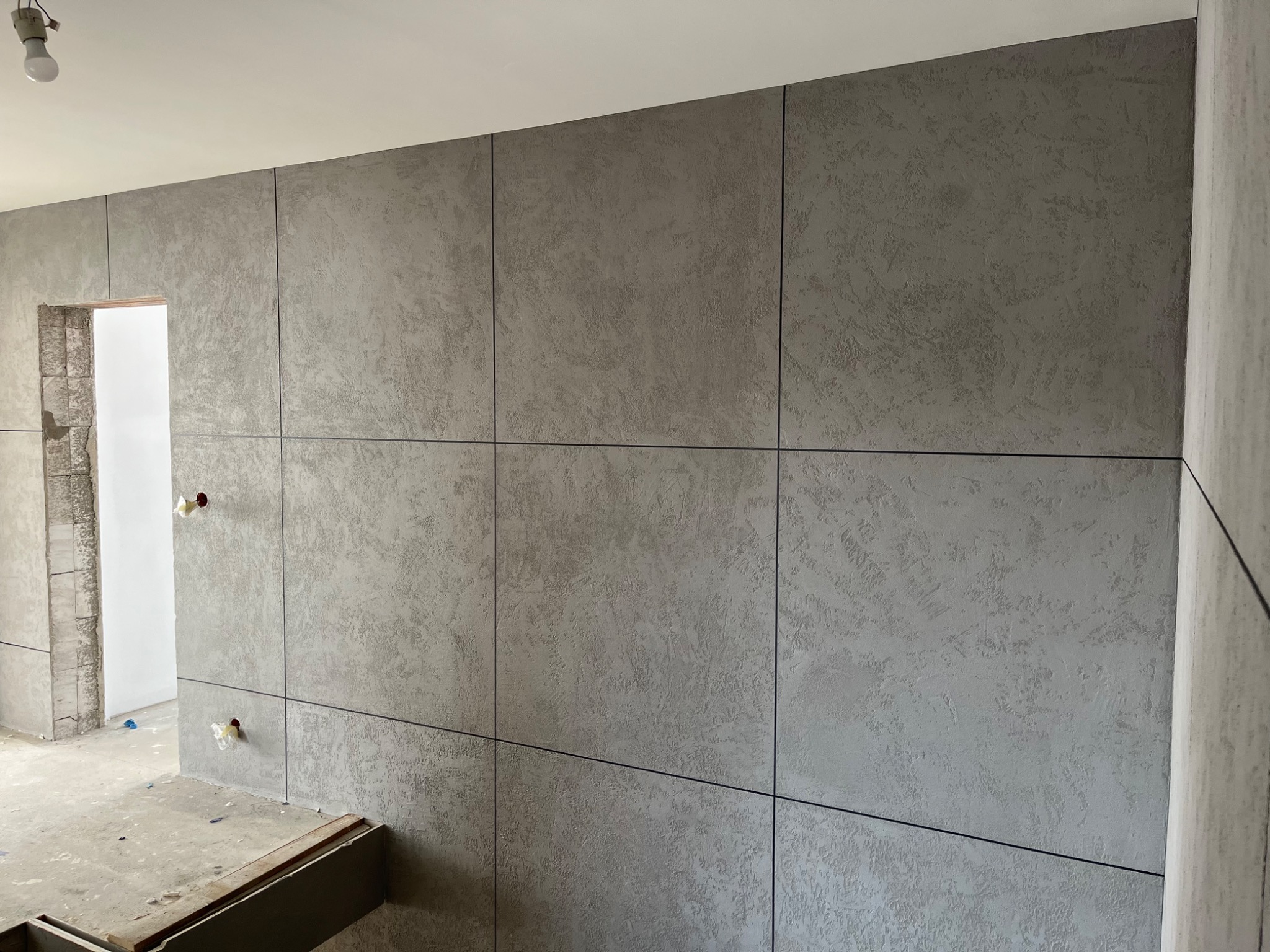 Ściana pokryta szarym tynkiem dekoracyjnym podzielona na panele, widoczne puszki instalacyjne zabezpieczone folią, surowy stan wykończenia pomieszczenia.