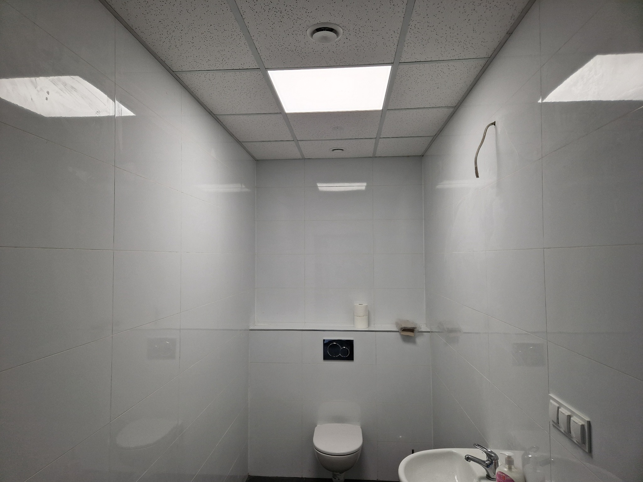 Wnętrze wykończonej łazienki z białymi, błyszczącymi płytkami, podwieszaną toaletą i umywalką z baterią oraz dozownikiem mydła. Na ścianie widoczne przełączniki elektryczne i wystający kabel.