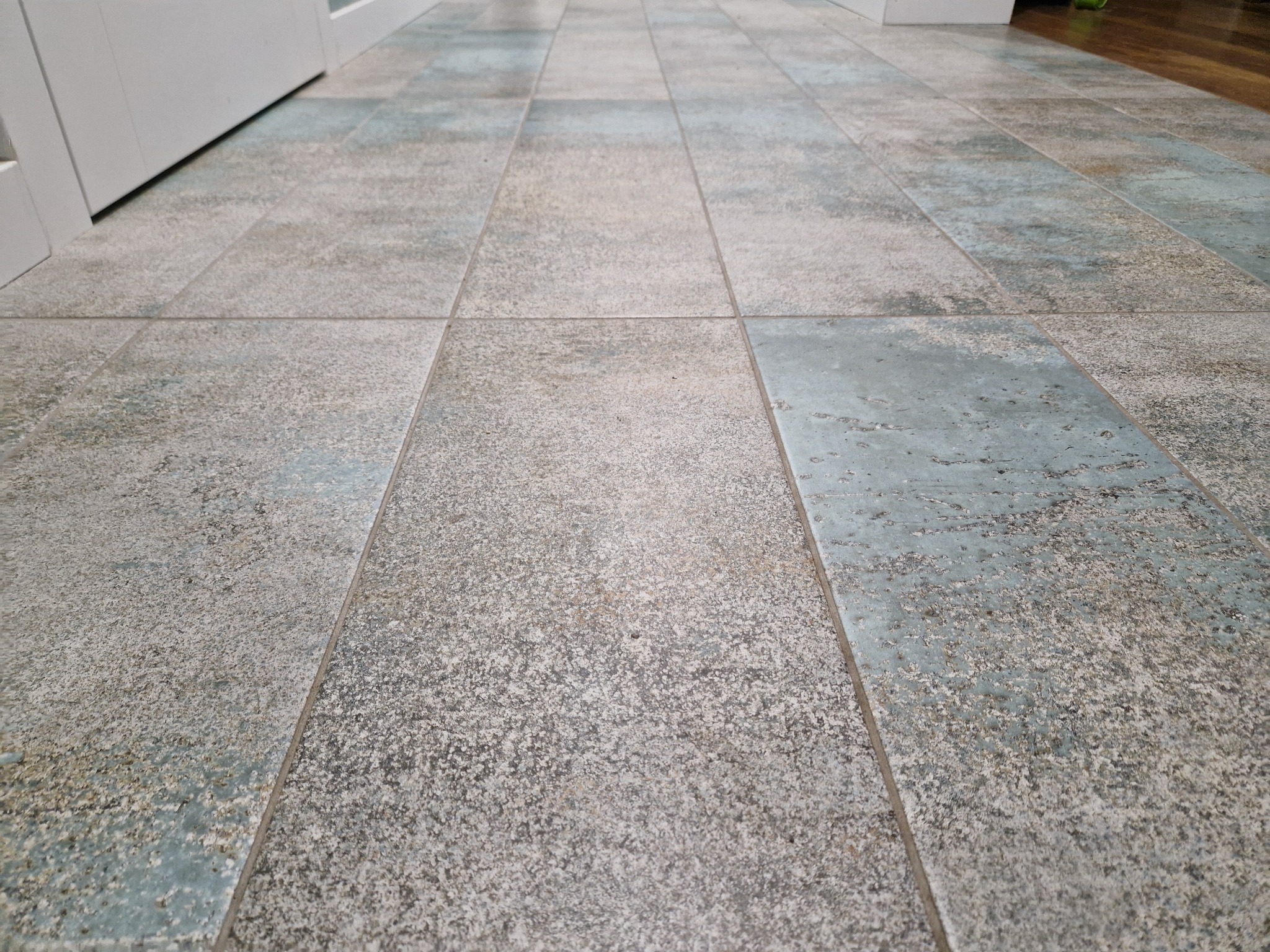 Ułożona podłoga z płytek ceramicznych w odcieniach szarości i błękitu, widoczne fugi i fragment białych drzwi w tle.